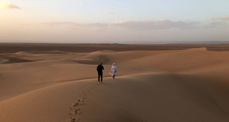 Two travellers walking along a dune ridge at dusk with footprints trailing behind.