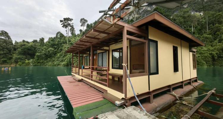 Bungalow jaune flottant ancré sur une eau émeraude face à une dense jungle en montagne.