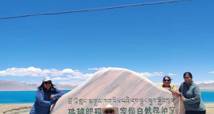 Des voyageurs sourient à côté d'une grande pancarte en pierre ornée d'inscriptions tibétaines surplombant un lac d'un turquoise éclatant.