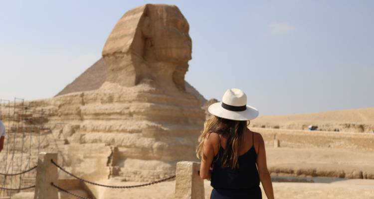 Woman in hat observing the Great Sphinx from behind a barrier on a sunny day.