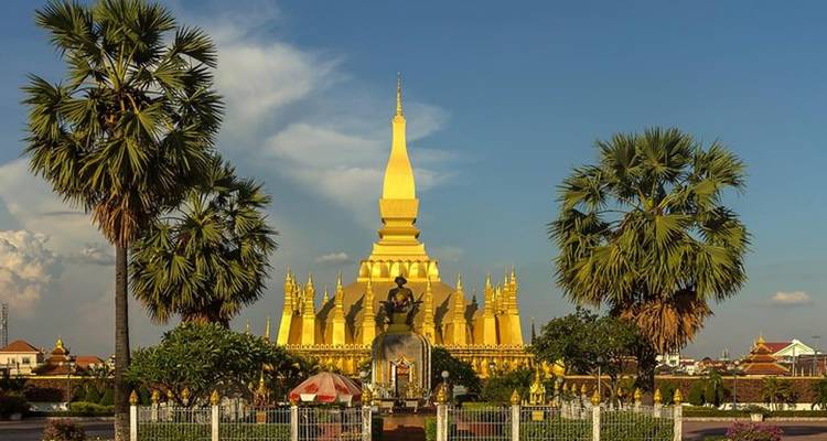 Golden Pha That Luang stupa flanked by palm trees shining against a clear blue sky.