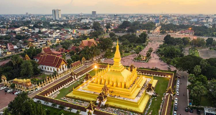 Drone view of Pha That Luang glowing at dusk, surrounded by Vientiane cityscape lights.