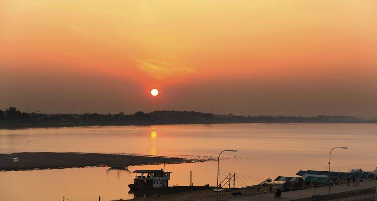 Orange sun setting over a calm Mekong River with boats and sandy banks in silhouette.