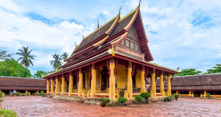 Ornate Lao Buddhist temple with tiered roof and golden pillars set around a brick courtyard on a bright day