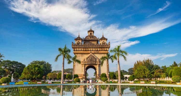 Patuxai monument framed by palm trees and blue sky with reflection in pond at midday