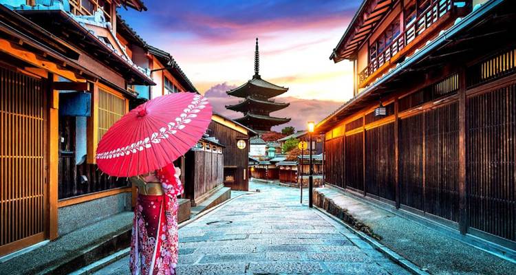 Kimono-clad woman with red parasol walks down Yasaka street toward the iconic wooden pagoda at sunset