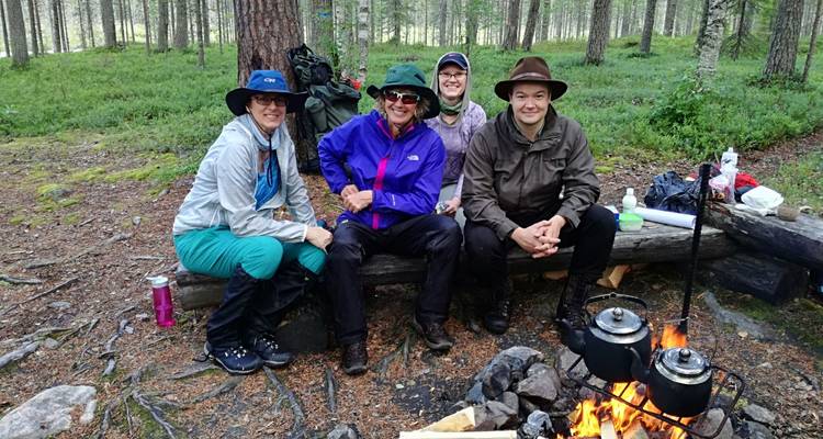 Hikers in outdoor gear smile beside campfire and kettles in Finnish pine forest