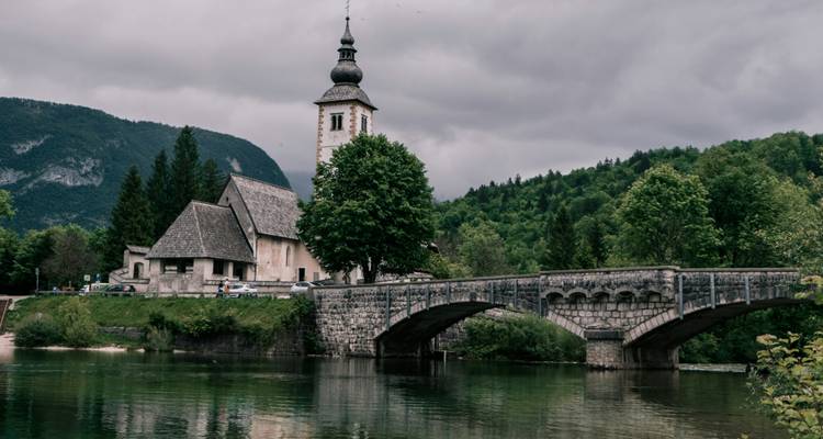 Historische kerk en stenen brug over smaragdgroene rivier bij Meer Bohinj op een bewolkte dag.