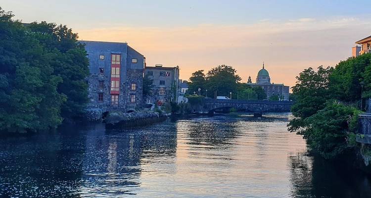 Scène fluviale sereine à Galway au coucher du soleil avec un pont en pierre et un dôme de cathédrale à l'horizon.