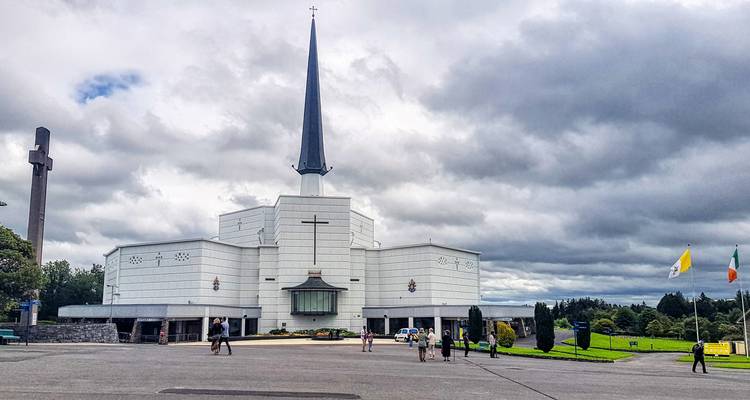 Basilique moderne du sanctuaire de Knock avec un haut clocher blanc sous des nuages gris dramatiques.