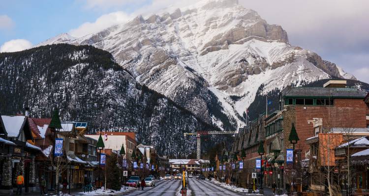 Schneebedeckte Hauptstraße von Banff, die zum aufragenden Cascade Mountain unter einem bewölkten Winterhimmel führt.