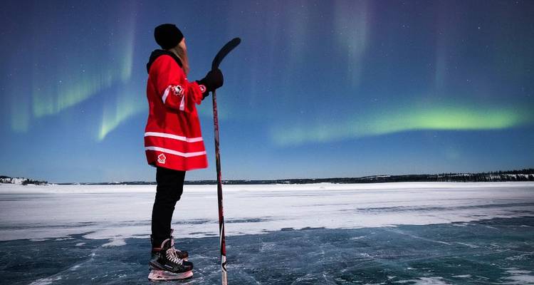 Eine Person in einem roten Eishockey-Trikot steht auf einem gefrorenen See mit Schlittschuhen und Schläger unter dem leuchtenden Polarlicht.