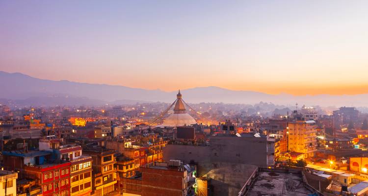 Kathmandu-Skyline bei Sonnenaufgang mit der weißen Boudhanath-Stupa, die über farbenfrohen Gebäuden und violetten Bergen aufragt.