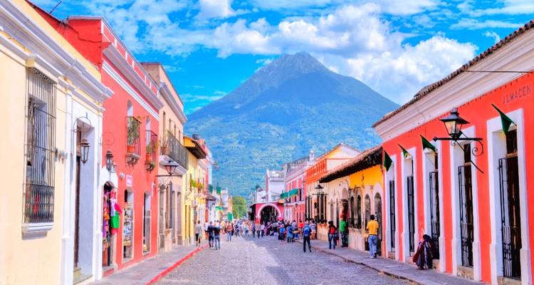 Calle de adoquines llena de vida bordeada de coloridos edificios coloniales que conduce hacia un imponente volcán.