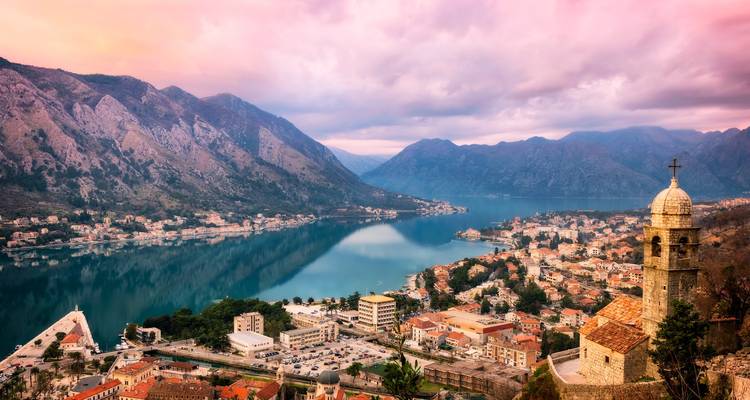 Erhöhter Ausblick über die Bucht von Kotor mit Spiegelung der umliegenden Berge und Terrakotta-Dächer unter rosa Himmel.