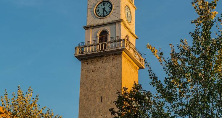 Steinerner Glockenturm, der in warmes Abendlicht getaucht und von grünen Blättern umrahmt wird.