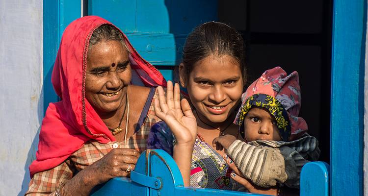 Una abuela sonriente, una madre y un bebé saludan desde la ventana de una casa azul brillante.
