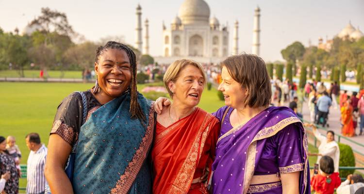 Drei Frauen in leuchtenden Saris posieren glücklich, während das ikonische weisse Marmor-Taj Mahal hinter ihnen im sanften Nachmittagslicht aufragt.