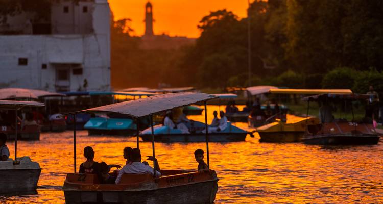Kleine Paddelboote mit Passagieren auf einem orange beleuchteten See bei Sonnenuntergang mit Silhouette eines Turms im Hintergrund