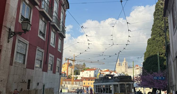 Calle histórica de Lisboa con un tranvía vintage, fachadas de edificios color carmesí y cables aéreos contra un cielo dramático.