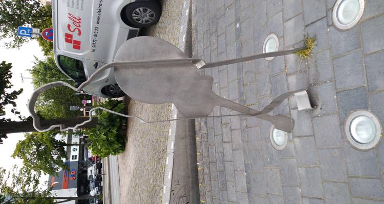 Sculpture en métal fin d'un bassiste sur un trottoir avec une camionnette garée et de la verdure à proximité