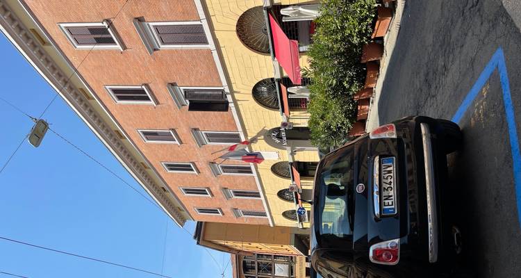 Scène de rue avec une petite Fiat noire garée devant un bâtiment d'hôtel traditionnel en briques et stuc