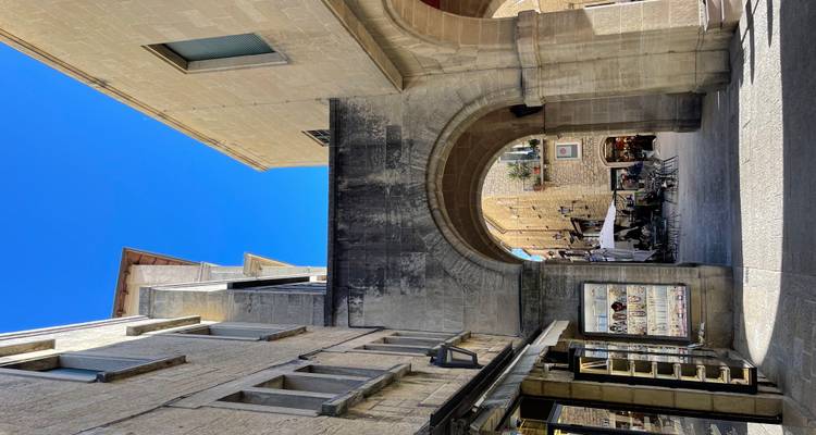 Passage voûté en pierre menant à un petit espace café dans une ville historique fortifiée sous un ciel bleu éclatant
