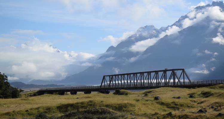 Un pont à treillis d'acier enjambe une plaine herbeuse avec des montagnes enveloppées de nuages se dressant à l'arrière-plan.