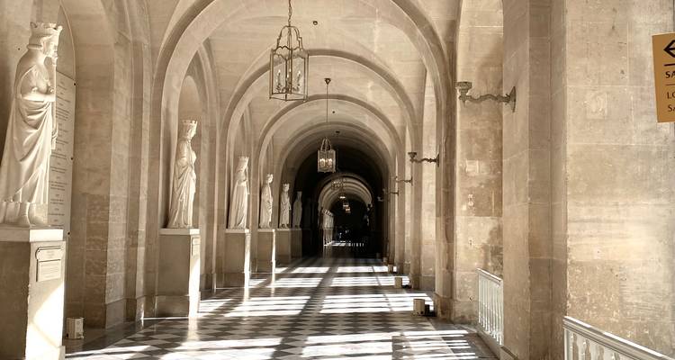 La lumière du soleil traverse les colonnades en arc de cercle bordées de statues classiques le long d'un corridor aux carreaux en damier.