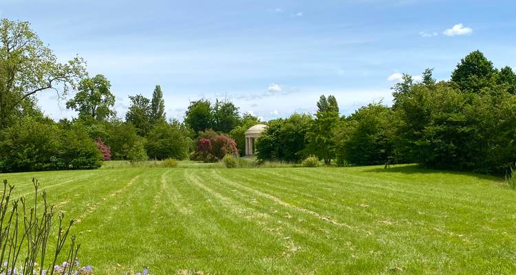 Une vaste prairie verdoyante parsemée d'arbres mène à un pavillon classique lointain sous un ciel bleu.