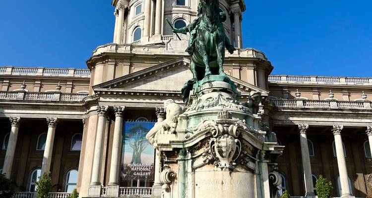 Bronzene Reiterstatue vor der prächtigen Fassade der Burg Buda unter einem klaren blauen Himmel.