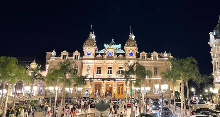 Illuminated Monte Carlo Casino façade at night with palm trees, bustling crowd and parked luxury cars.