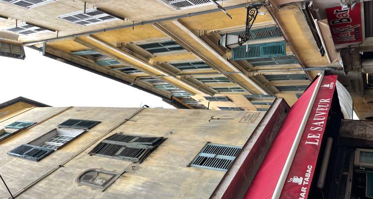 Narrow Mediterranean alley with tall pastel buildings, green shutters and a red café awning.