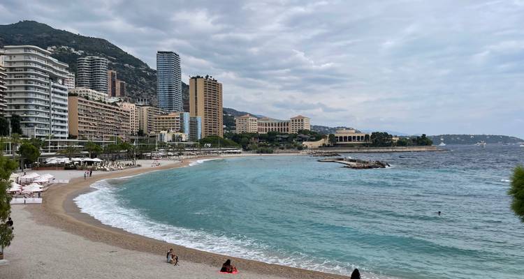Curving sandy beach with turquoise water, sunbathers and Monaco skyline of tall hotels by the sea.