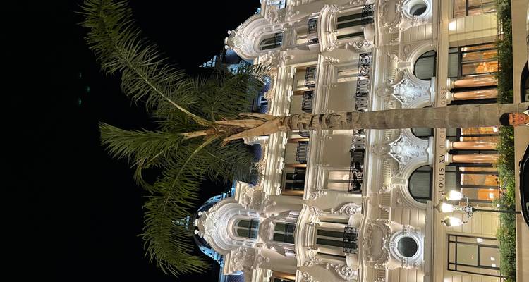 Nighttime shot of an ornate Belle Époque hotel façade with lit balconies and a tall palm tree in front.