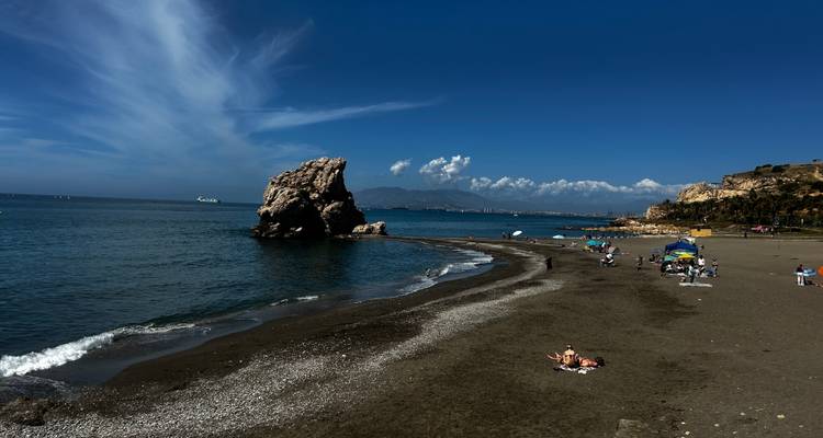A quiet beach with a solitary rocky stack offshore; sunbathers and umbrellas dot the dark sand beneath a vivid blue sky.