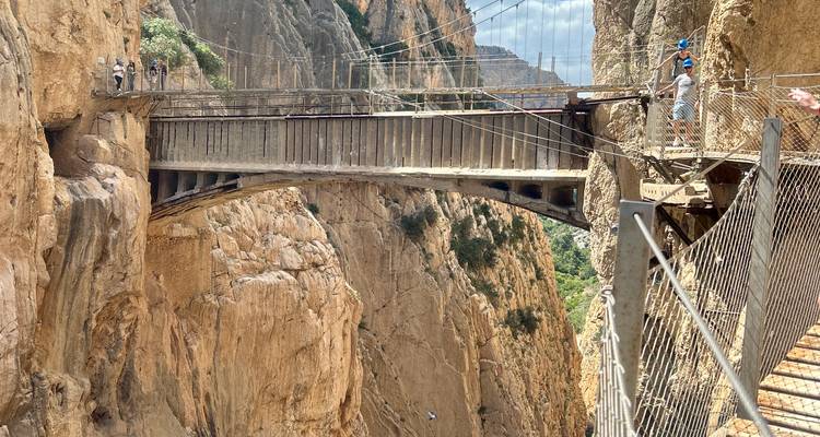 Visitors traverse the suspended bridge of the Caminito del Rey between sheer orange canyon walls.