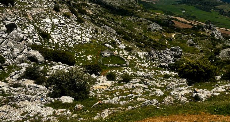 Rock-strewn slopes descend to a small grassy basin with winding paths across a patchwork of green fields.