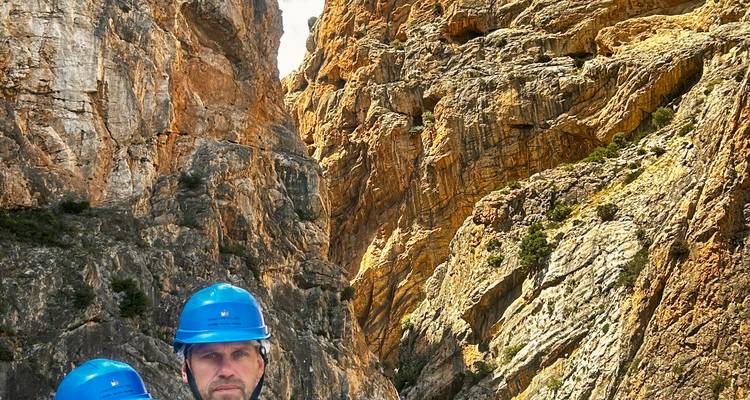 Two helmeted visitors stand close to the camera with towering orange canyon walls behind them.