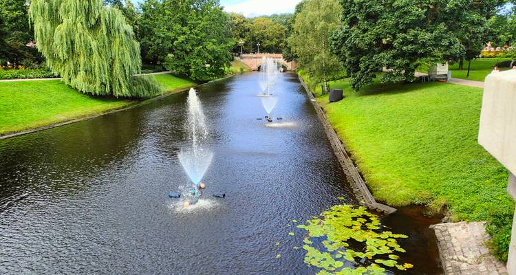 Canal with water fountains and water lilies bordered by green lawns and trees.
