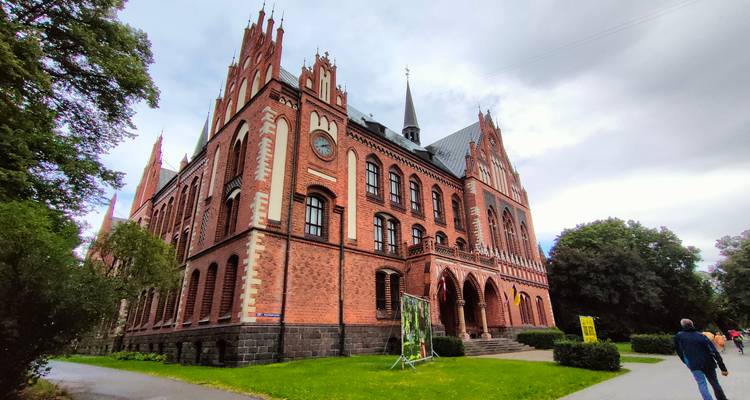 Large red-brick neo-Gothic building with turrets and arched windows along a tree-lined path.
