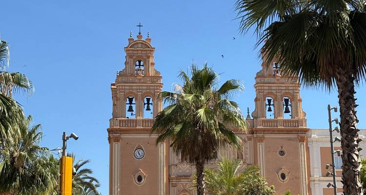 Façade d'église baroque à deux tours avec des palmiers sur un ciel bleu dégagé.