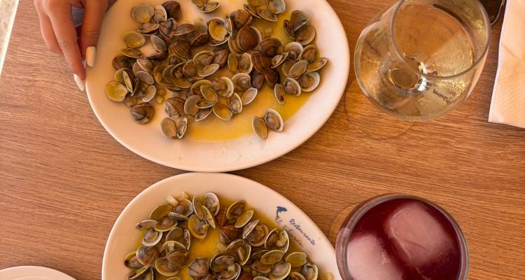 Tapas de fruits de mer sur table avec des assiettes de palourdes, des verres de vin et la main d'un client qui se tend vers les plats.