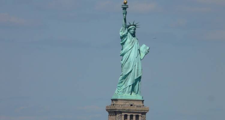 Close-up telephoto shot of the Statue of Liberty’s upper half against a pale blue sky.