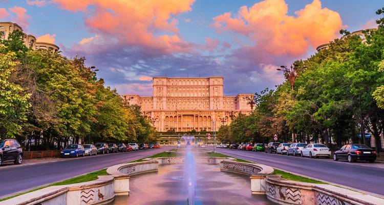 El monumental Palacio del Parlamento en Bucarest enmarcado por un cielo de atardecer en tonos pastel y un bulevar bordeado de fuentes.