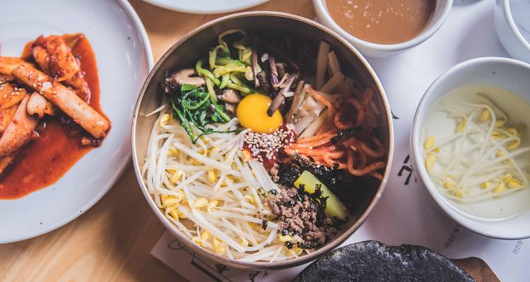Un bol coloré de bibimbap entouré de plats d'accompagnement coréens sur une table en bois.
