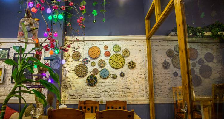 Farbenfrohe, eklektische Café-Innenausstattung mit hängenden Laternen, gewebter Tellerkunst an einer weißen Ziegelmauer und Holztischen