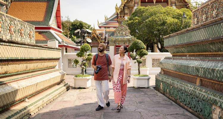 Young couple strolling between ornately tiled walls and pagodas in a Bangkok temple complex.