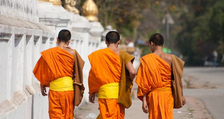 Three novice monks in orange robes walk along a temple wall carrying alms bags.