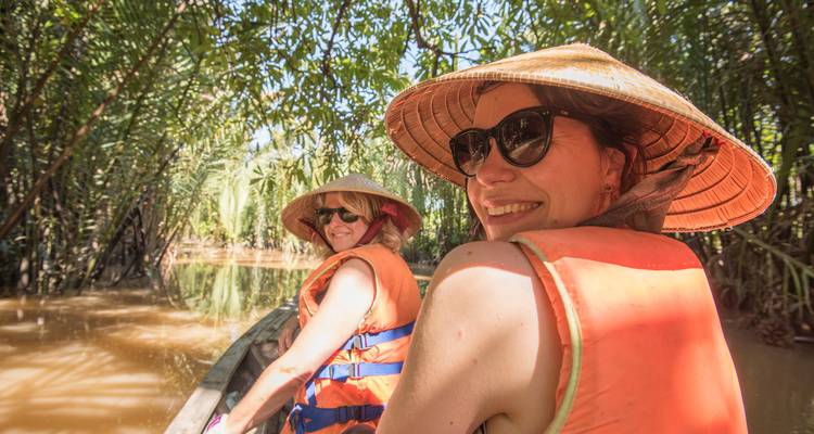 Travellers in life vests and straw hats smile while paddling a boat through palm-lined brown waterways.
